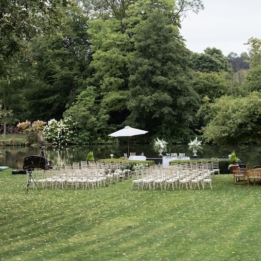 White chairs are arranged in rows on a grassy lawn for an outdoor event, surrounded by green trees. A few tables, umbrellas, and benches are also set up, with a river and dense foliage in the background.