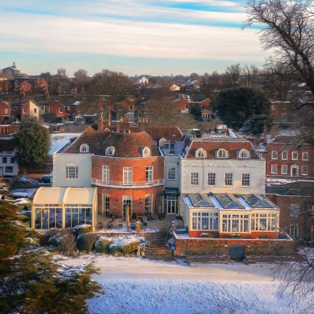 Large red and white mansion with glass extensions in a snowy, suburban neighborhood, surrounded by trees and smaller brick houses under a partly cloudy sky.